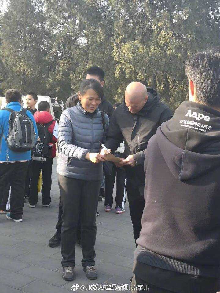 在清晨柔和的光线下，足坛传奇齐达内身穿简约的深色外套，如同一位普通游客漫步在北京天坛，他标志性的光头在古老松柏的映衬下格外醒目，脸上带着轻松惬意的微笑。