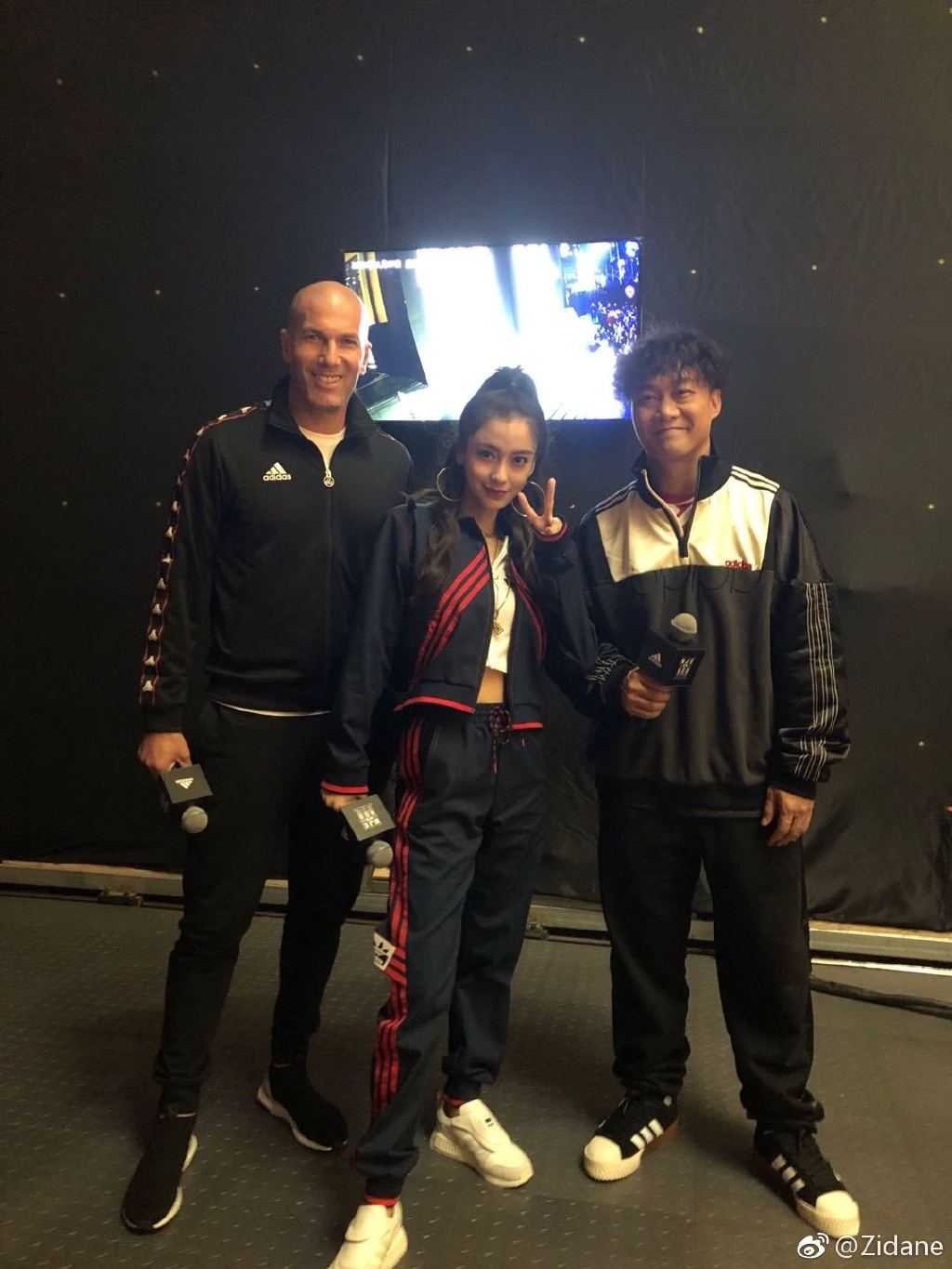 齐达内与陈奕迅和Angelababy同框，三人手持乒乓球拍，对着镜头露出灿烂笑容，背景是热闹的品牌活动现场。