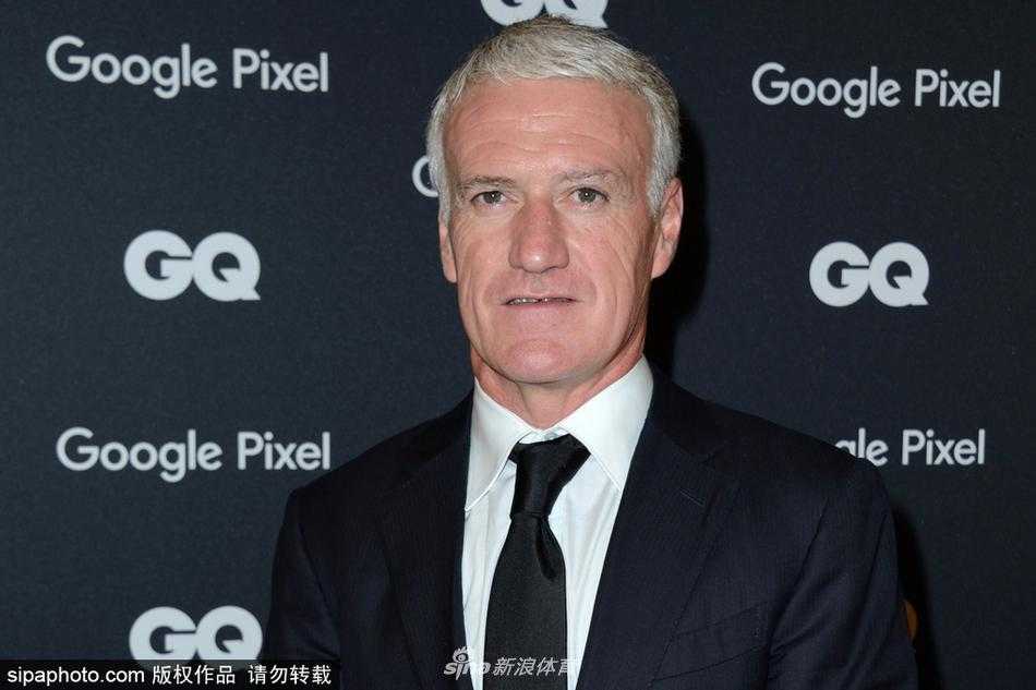 德尚在颁奖礼上手持奖杯与人合影，脸上洋溢着谦逊而满足的微笑，背景是典雅的室内灯光，突显其平易近人的一面