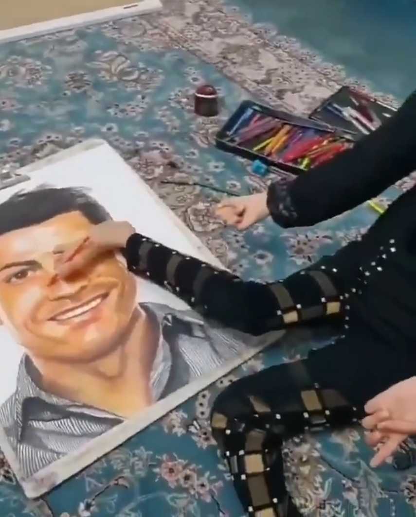 法特梅正全神贯注地用脚趾夹着画笔，在画板上细细描绘C罗的轮廓，眼神中充满坚定与热爱。