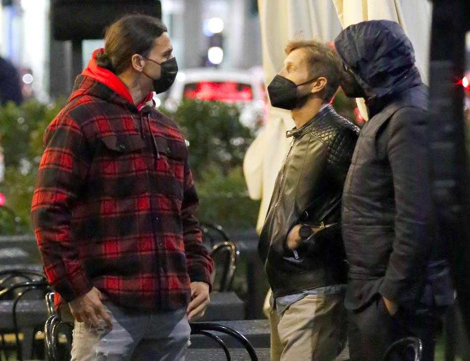 又一位球迷加入合影行列，伊布依然保持着标志性的挺拔站姿，耐心配合。餐桌上摆放着未喝完的饮品，背景里餐厅的装潢古朴雅致，仿佛在诉说着米兰城独有的足球文化与生活节奏。