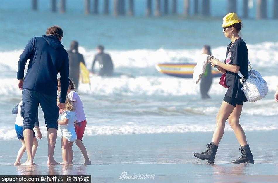 布莱德利·库珀赤脚走在沙滩上，温柔地抱着女儿，伊莉娜跟在身后，一家三口画面温馨和谐。