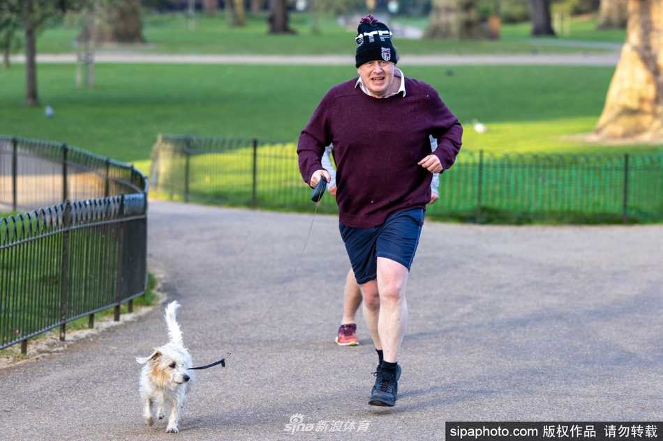 英国前首相鲍里斯·约翰逊身穿深色运动装，头戴一顶柔软的蓝色毛线帽，在晨光中牵着活泼的爱犬迪林慢跑。他的脸上带着轻松的微笑，眼神随和地望向前方，步伐稳健，展现出工作之外充满活力与亲和力的一面。