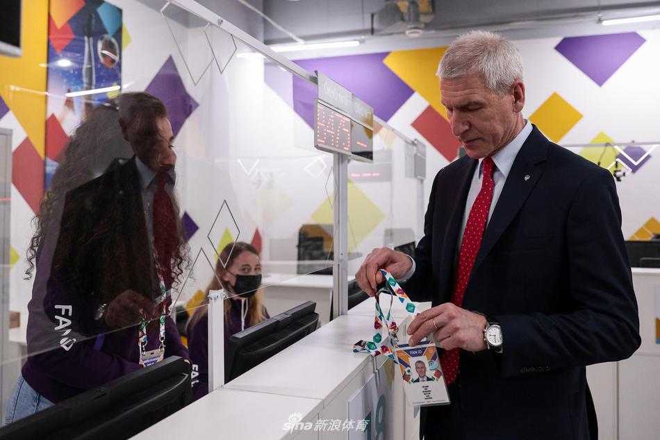 马特钦部长低垂眼帘，神情专注地翻阅着手中的球迷证文件册，背后的场馆通道灯光昏黄，营造出一种赛前特有的静谧与严肃氛围，仿佛在审阅的不是一张证件，而是整个赛事的组织流程。