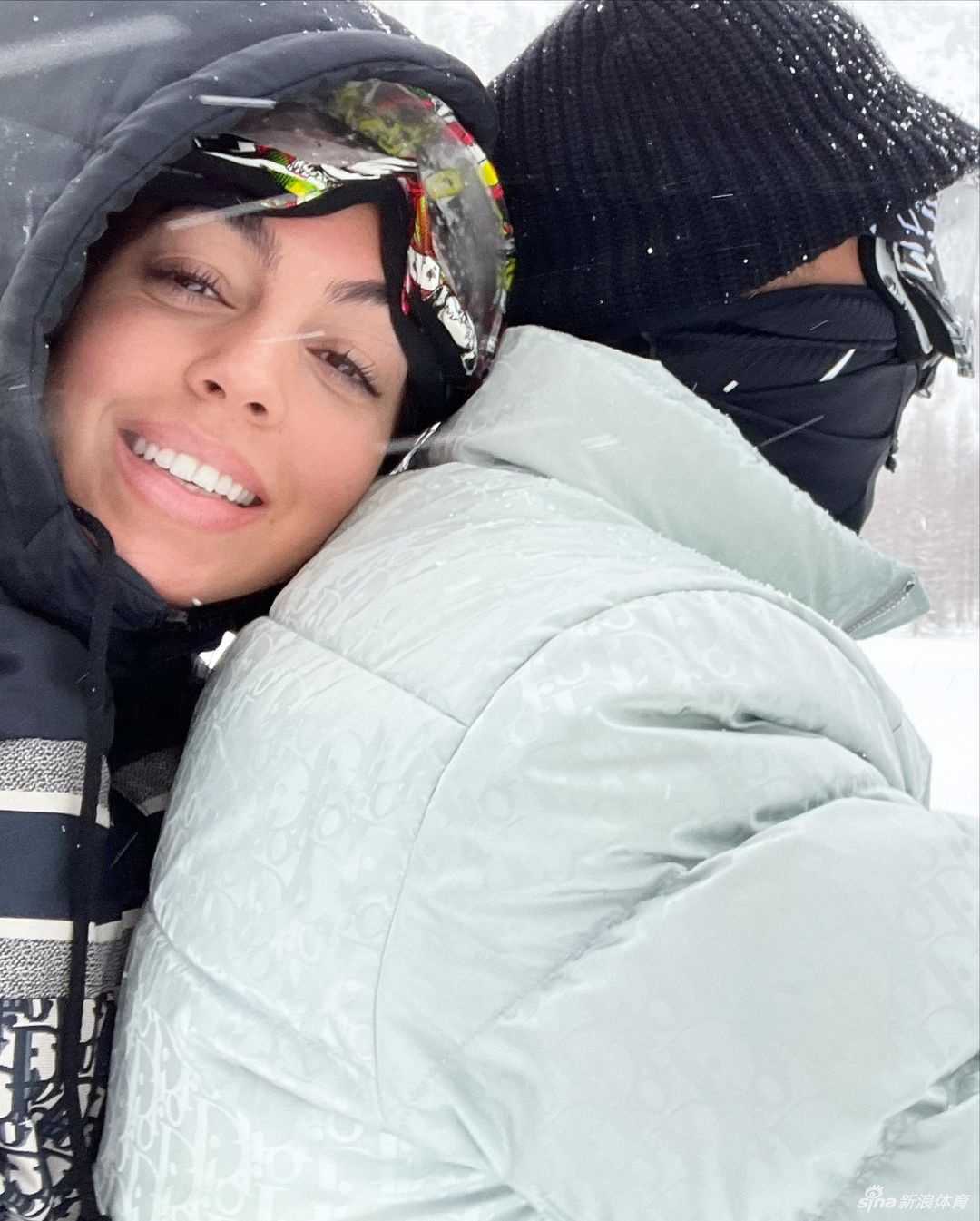 温馨的亲子时刻，C罗坐在雪地上，怀里抱着可爱的小女儿阿拉娜·玛蒂娜。父亲眼神温柔地注视着女儿，阿拉娜则好奇地望向镜头，父女间的互动充满了宠溺与爱意，一旁的雪地摩托和远处的雪山构成了这幅幸福图景的背景。
