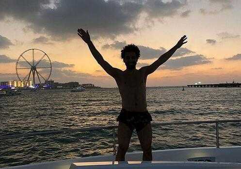 利物浦球星萨拉赫戴着墨镜半裸上身，露出健硕肌肉，惬意地躺在度假酒店的躺椅上享受日光浴，脸上洋溢着轻松的笑容。