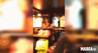 在巴萨商店巨大的梅西海报前，两名利物浦球迷笑容满面地配合着，其中一人将手中的气球欧冠奖杯高高举过头顶，对准海报上的梅西，仿佛正在向海报中的巴萨球星展示这座他们刚赢得的冠军奖杯，画面充满戏剧感和挑衅意味