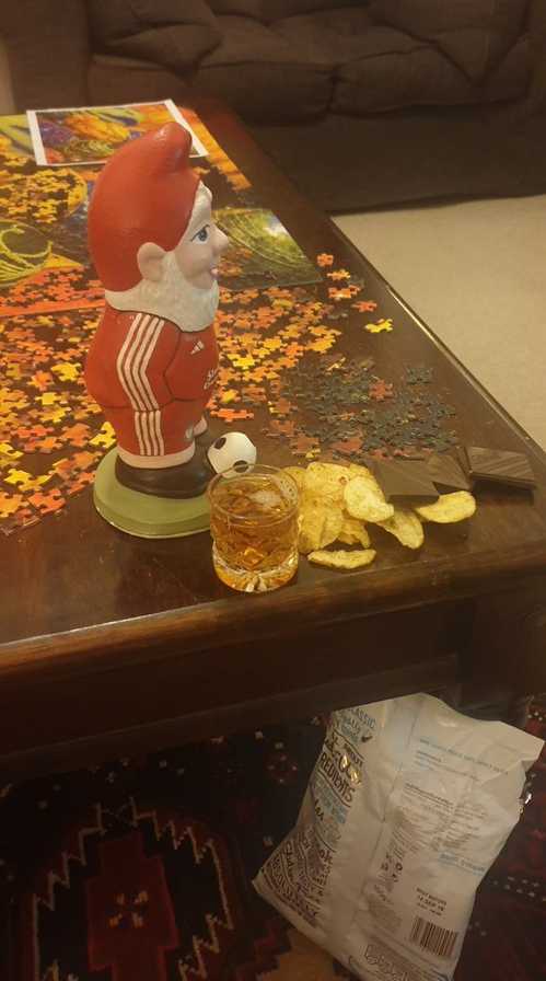 一张充满岁月感的照片里，红衣小矮人被放在安菲尔德球场看台的栏杆上，背景是KOP看台掀起的红色人浪，它渺小却醒目，像一位沉默的场上第十二人，静静凝视着绿茵场上的英雄们。