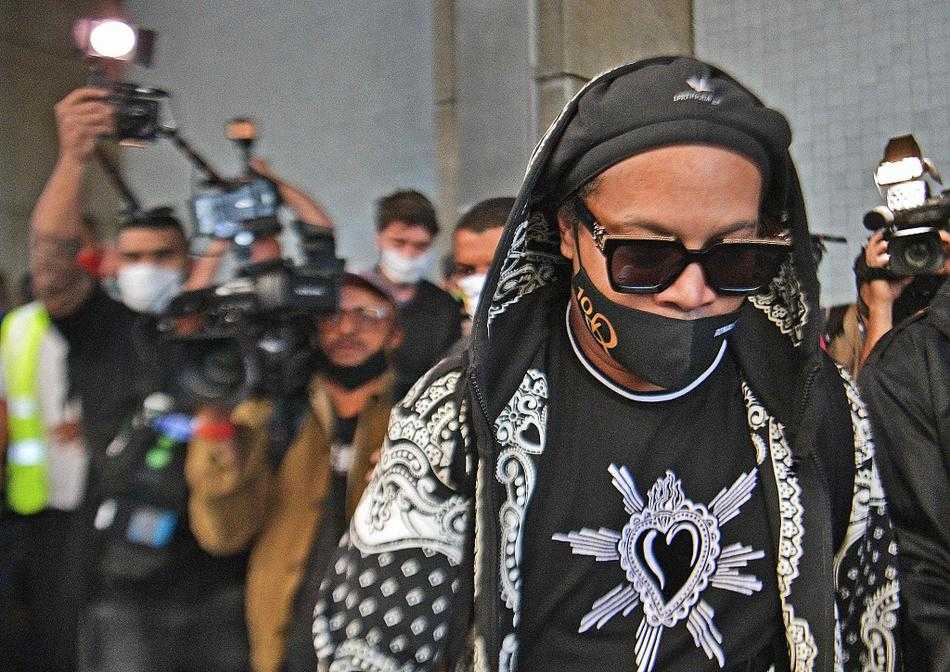 小罗的哥哥阿西斯紧随其后，神情严肃地拎着行李袋。兄弟二人共同经历了这场风波，此刻在人群中并肩而行，准备一同离开。