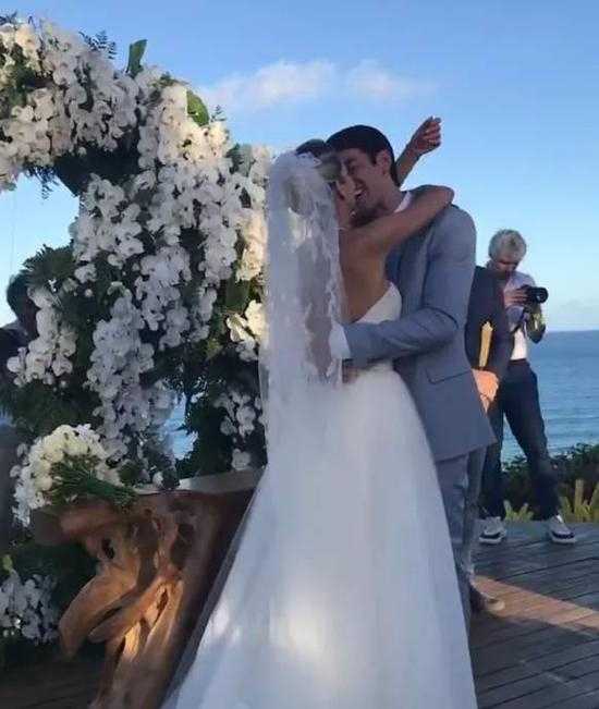 卡卡牵着妻子卡罗尔的手走在鲜花拱门下，新郎一身浅灰色西装英俊挺拔，新娘的拖地头纱在微风中轻扬，画面唯美而神圣