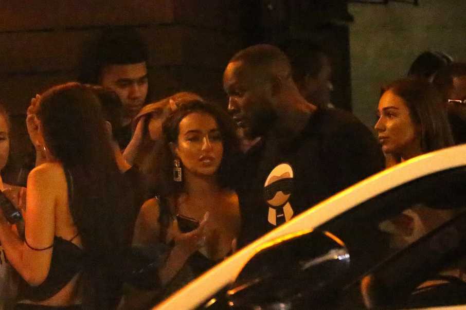 卢卡库身穿休闲T恤，在洛杉矶夜店的柔光下与一位神秘女子凑近交谈，他脸上挂着孩子般的笑容，女子则用手掩嘴轻笑，氛围轻松而亲密。