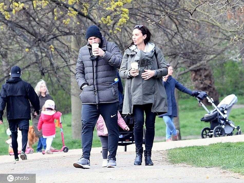 妻子克里斯汀身穿休闲灰色卫衣，用婴儿背带将小弗雷迪紧紧抱在胸前，一家人在公园小径上悠闲前行，画面充满了宁静与幸福的气息。