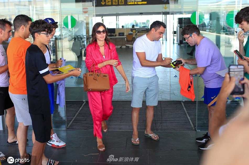 哈维与妻子努莉亚在巴塞罗那机场大厅并肩而行，两人面带轻松笑容，哈维身着休闲西装，妻子挽着他的手臂，展现夫妇间的温馨默契。