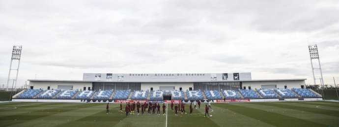 国足集训队全体教练员与球员在宏伟的皇马体育城入口处合影留念，背后的队徽闪耀着欧冠冠军的光芒，每个人的脸上都写满了对这次顶级交流之旅的期待与专注。