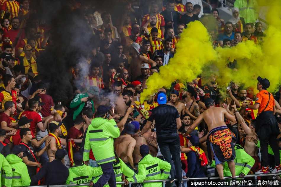 一群身着白色球衣的阿尔萨德球迷面色严峻地站在看台栏杆后，他们的眼神中透露出对球队大比分落后的失望以及对看台另一侧对手球迷举动的警惕。