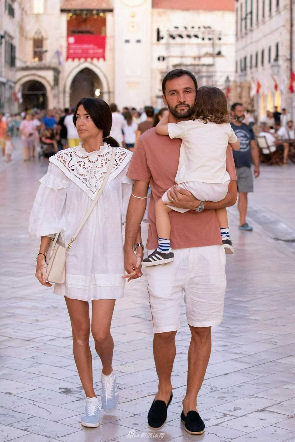 夏日的杜布罗夫尼克古巷中，巴德利双手稳稳地环抱在女儿胸前，将宝贝轻柔地护在怀中，妻子伴随身旁，一家三口在斑驳的墙影下悠然前行，仿佛一幅流动的温馨画卷。