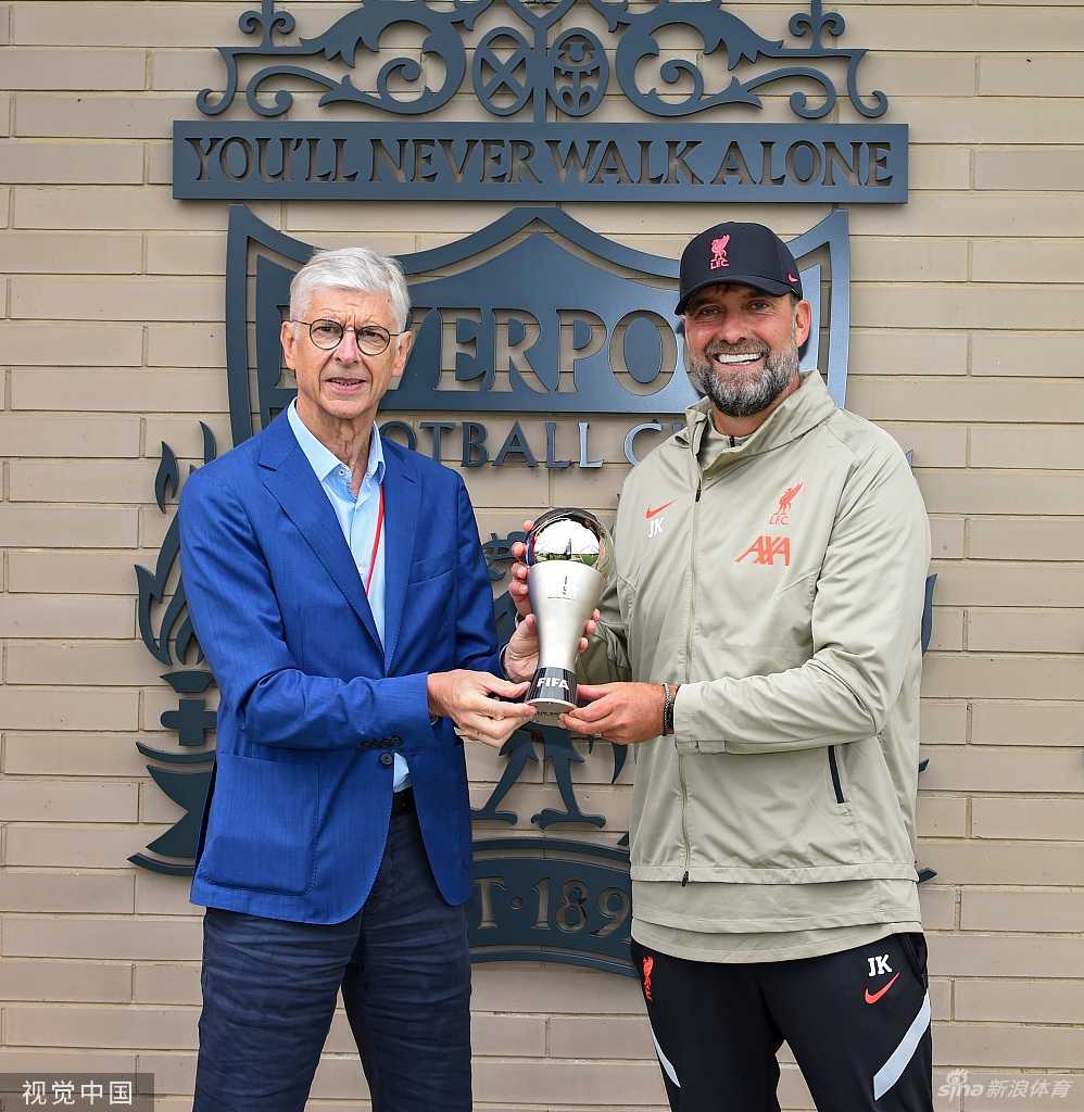 画面聚焦于克洛普手捧FIFA年度最佳主教练奖杯，他标志性的络腮胡和灿烂笑容融为一体，眼中闪烁着激动的光芒，身后的背景板印着利物浦队徽，仿佛整个世界都在为他喝彩。