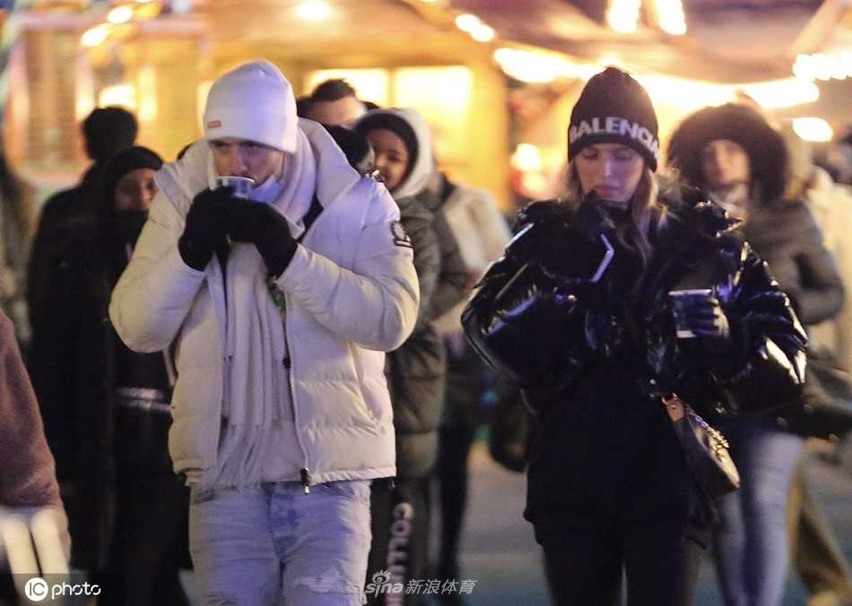 格里利什与女友萨莎在冬日乐园入口相视而笑，他戴着毛线帽，萨莎依偎在他身旁，背景是璀璨的灯光与欢乐的人群。