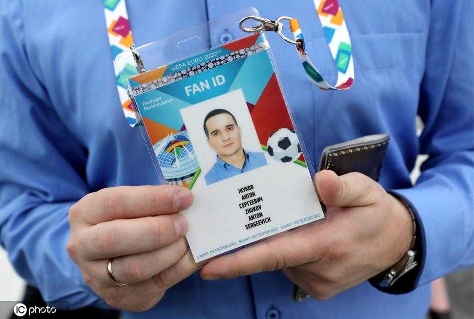 一名球迷正俯身在申请台前填写表格，手中握着的笔尖落在“FAN ID”申请表的个人信息栏上，周围是熙熙攘攘排队的人群，远处玻璃门外还能看到涅瓦大街上冬日特有的行人剪影。