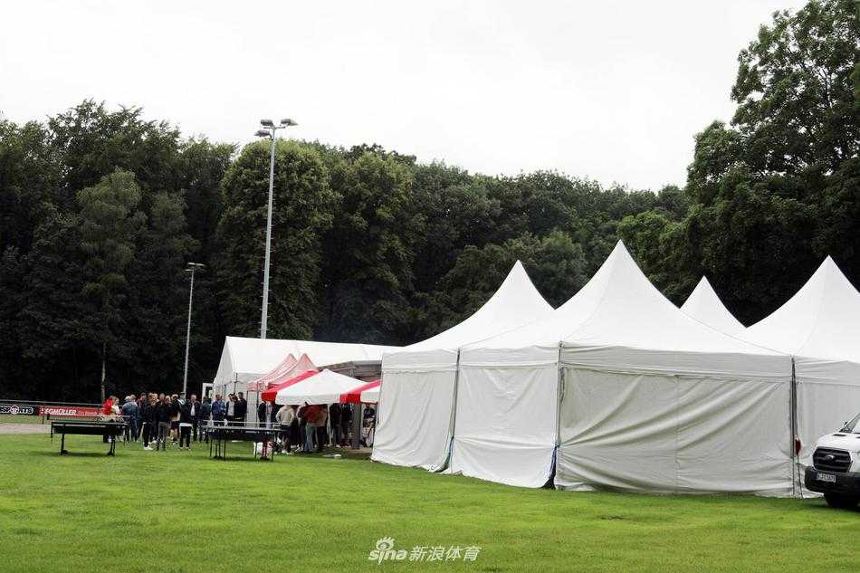 在夏日的阳光下，科隆俱乐部烧烤团建现场烟雾缭绕，一位教练正专注地翻转着烤架上的香肠和牛排，油脂滴落在炭火上发出滋滋响声，空气中弥漫着诱人的焦香