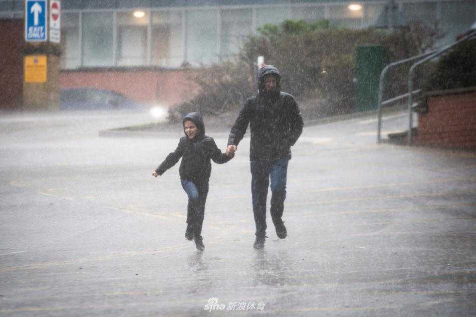 体育场外的台阶上积满了雨水，倒映着昏暗的灯光和几个匆匆而过的模糊身影。一对父子手牵手奋力往上走，父亲倾斜着身体为孩子挡住一部分斜吹过来的冷雨，小小的身影紧紧跟着父亲的步伐，这一幕让冰冷的石阶充满了温情。
