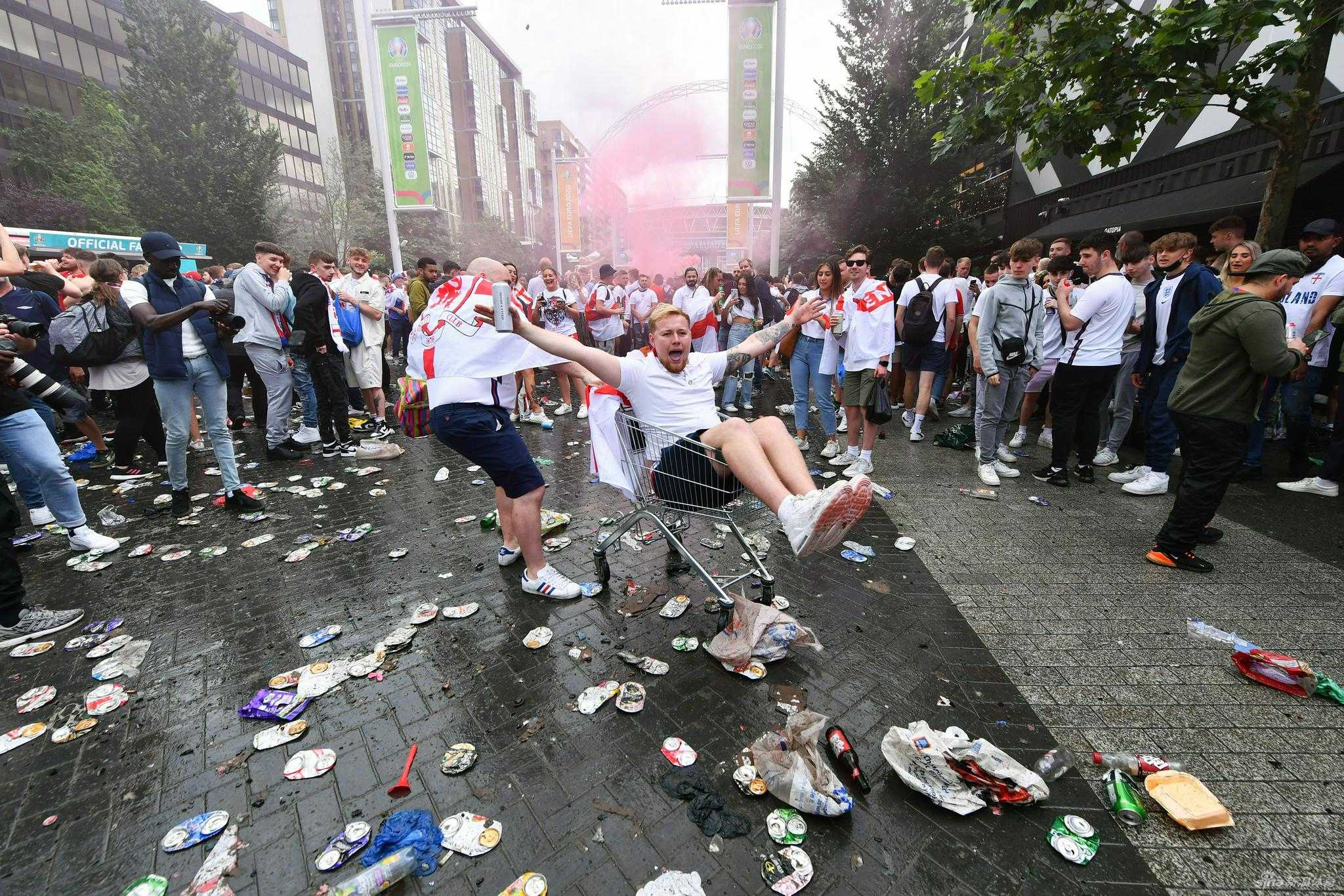 两名球迷在堆积如山的垃圾袋旁相拥，其中一人脸上画着圣乔治旗，神情中充满了失落与茫然。他们身后，清洁工忙碌的身影与这片狼藉形成鲜明对比，宣告着一场盛大的幻灭。