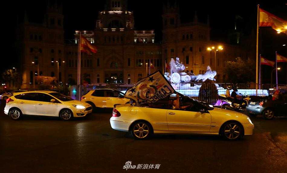 从高处俯瞰马德里市中心的主干道，夜色中亮着车灯的长龙缓缓前行，许多车辆的天窗或车窗中伸出白色的皇马队旗，旗帜在风中猎猎作响