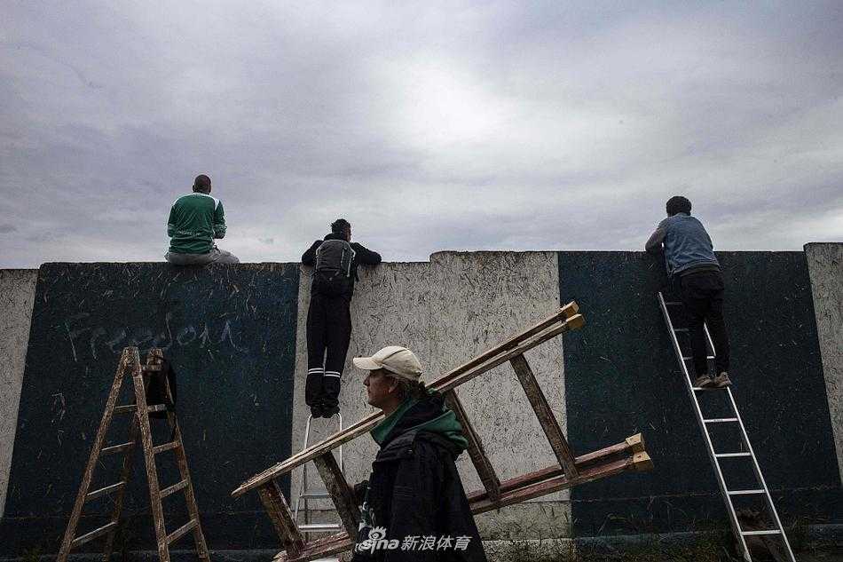 在捷克布拉格的一座球场外，两名身穿深色T恤的球迷为了看球，勇敢地攀爬在绿色的铁丝围栏上，他们的身影与围栏上印着的黄色联赛标志形成了鲜明对比，仿佛在诉说对绿茵场无尽的思念。