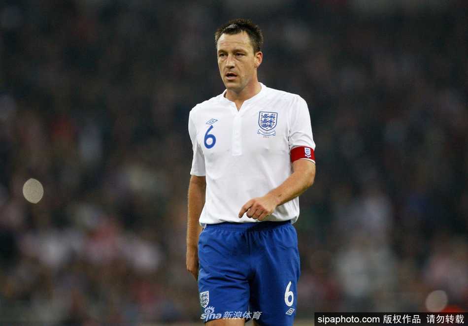 身披战袍的特里在比赛中神情专注，正大声指挥着队友防线，他手臂上的队长袖标在阳光下格外醒目，身后是看台上球迷挥舞的旗帜海洋