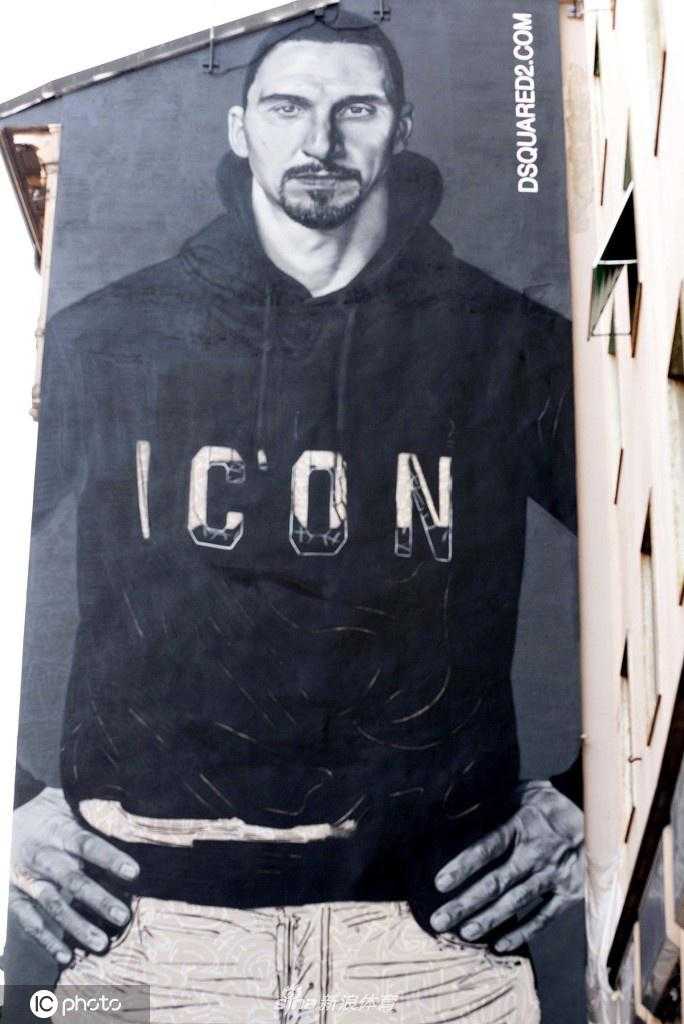 镜头拉远，展示了整面墙的壁画全貌。伊布双手自然下垂，身体微微前倾，姿态放松却充满内在张力。黑白灰的色调与周围建筑的红砖墙形成微妙的对比，巨大的尺幅让路过的行人在其脚下显得格外渺小，凸显了艺术作品的宏伟以及伊布作为城市英雄的崇高地位。
