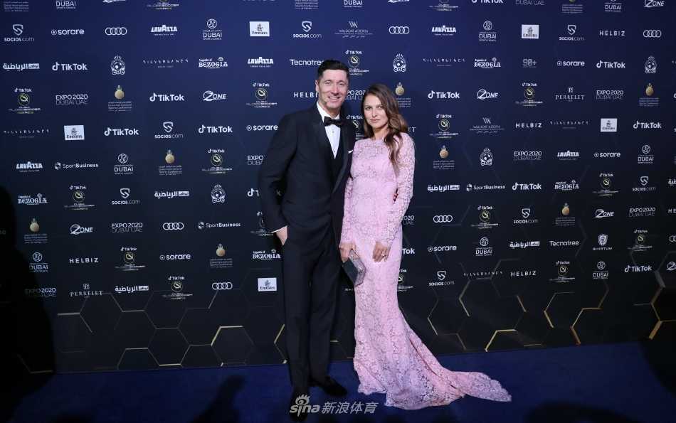 莱万多夫斯基身着经典黑色礼服，与身着一袭银色长裙的妻子安娜十指紧扣，在红毯聚光灯下绽放自信微笑，宛若好莱坞明星伴侣。