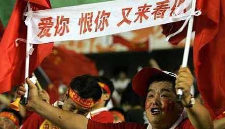 一位球迷高举着自制的硬纸板标语，上面画着一个戴着墨镜的卡通形象，旁边写着调侃国足长期表现不佳的幽默语句，引得周围观众会心一笑。