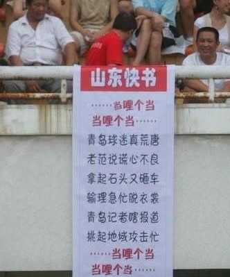 一名男球迷身披国旗，脸上画着油彩，双手高高举起一块写有谐音梗的创意标语牌，将国足球员的名字与网络流行语巧妙结合，趣味十足。