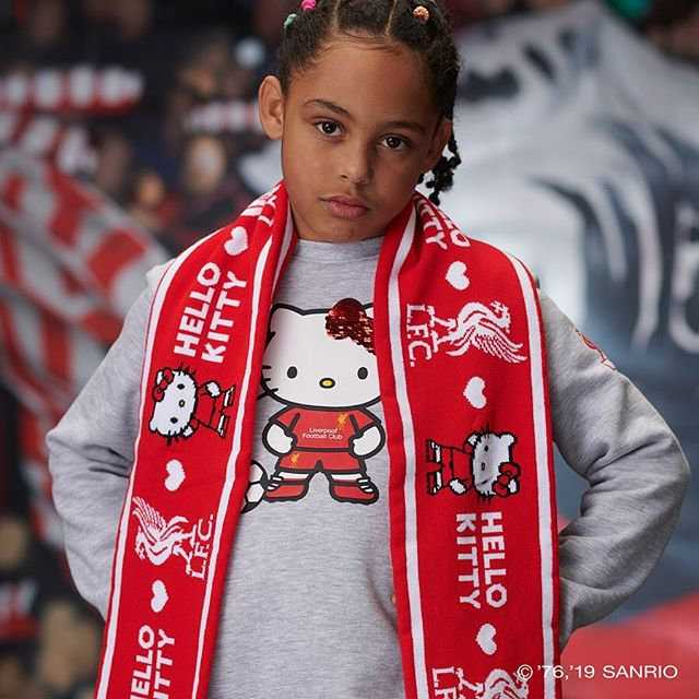一位小球迷身穿全套利物浦联名Hello Kitty的服装，开心地抱着新买的联名足球，脸上洋溢着纯真的笑容，身后是安菲尔德球场的留影。
