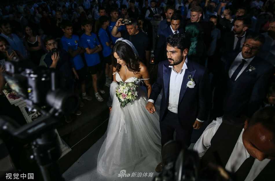 新婚夫妇在户外与众多亲友开心合影，人群簇拥着他们，背景是美丽的自然风光，共同分享着这份巨大的喜悦。
