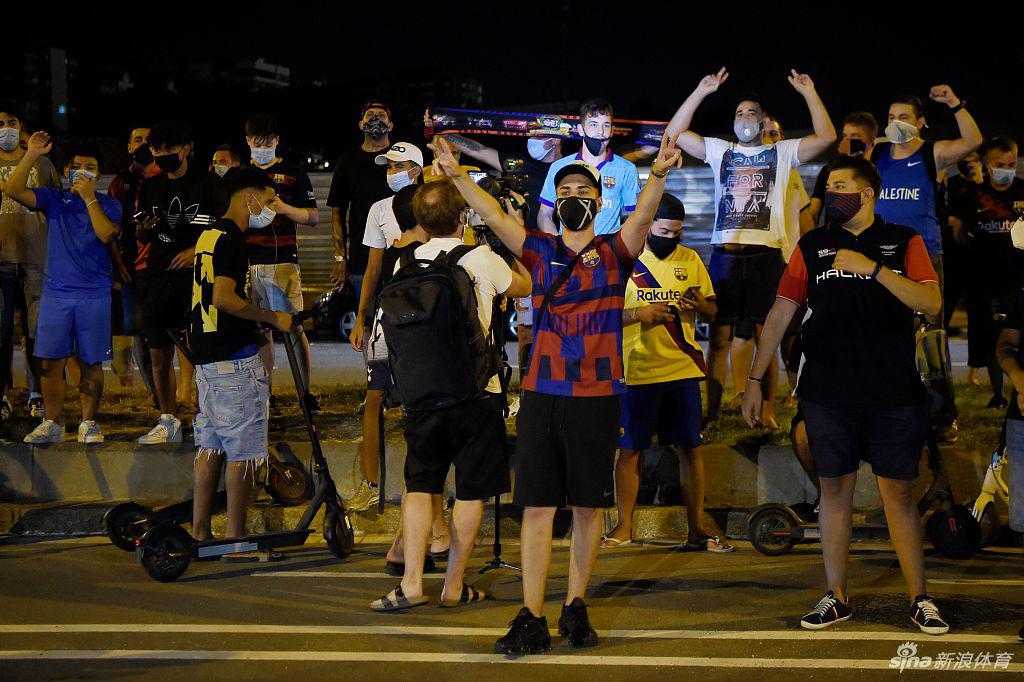 夜色渐浓，诺坎普球场外的灯光将聚集的人群身影拉长。几名年轻的球迷爬上了围栏，其中一人高高举起一面自制的巨大横幅，上面用醒目的字母写着对主席巴托梅乌的抗议标语，背景是模糊但情绪激动的密集人群。