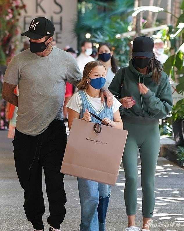 贝克汉姆一家三口行走在绿植环绕的街道上，贝克汉姆的手轻柔地搭在女儿小七的肩膀，目光温柔地看向前方，小七则拎着购物袋走在中间，画面充满了亲情的温暖与守护感。