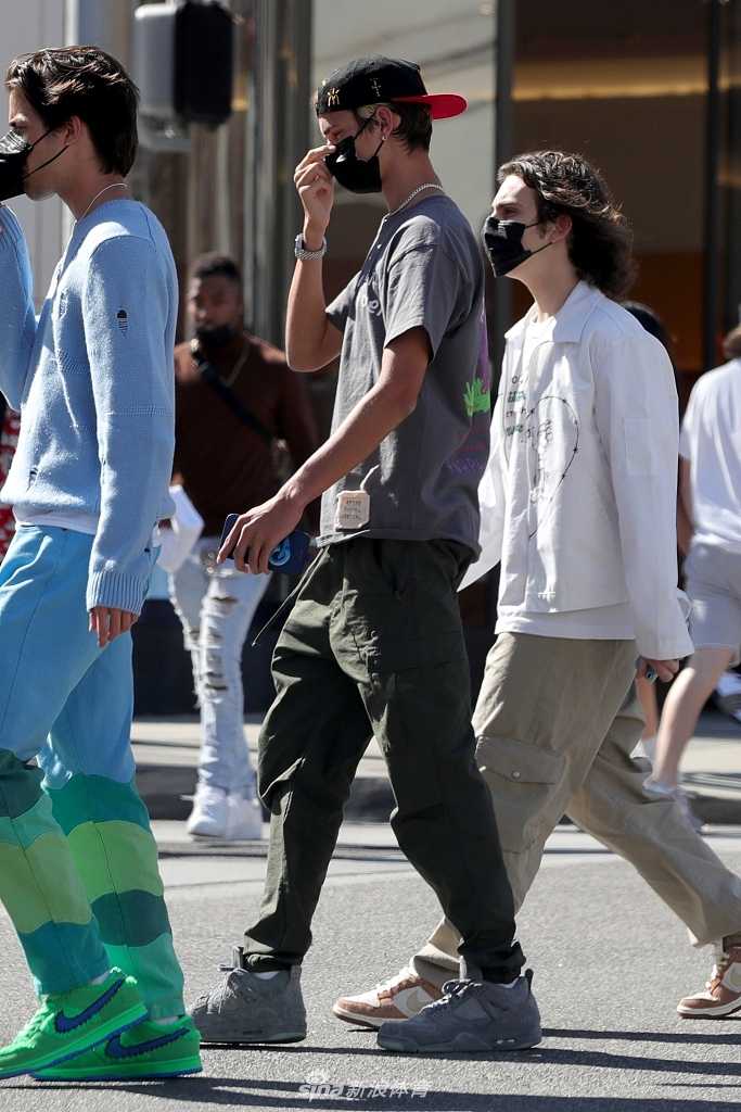 罗密欧反戴棒球帽走在洛杉矶街头，脸上洋溢着灿烂笑容，身后的高大保镖警觉地注视着四周，一黑一白的穿搭形成有趣对比。